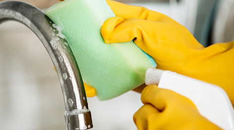 Picture of cleaning tap in sink with green sponge, yellow rubber gloves and cleaning spray