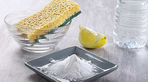 Photograph of cleaning supplies that include a sponge, bowl, lemon, bottle of water, and a plate of baking soda