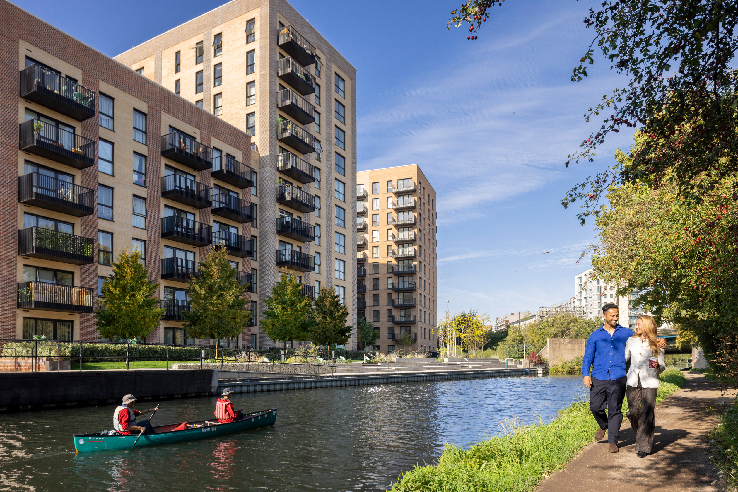 Two people walking down the canal with their arms around each other and two people in a canoe on the canal with the apartments in the distance