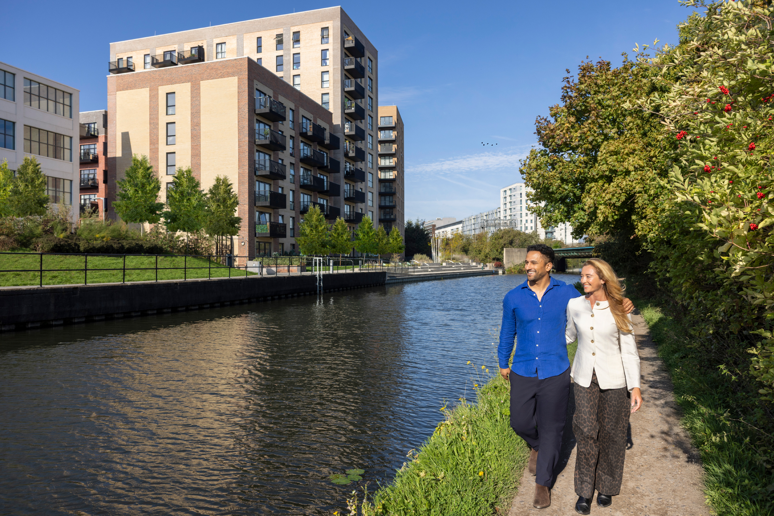 Two people walking down the canal with their arms around each other and the apartments in the distance